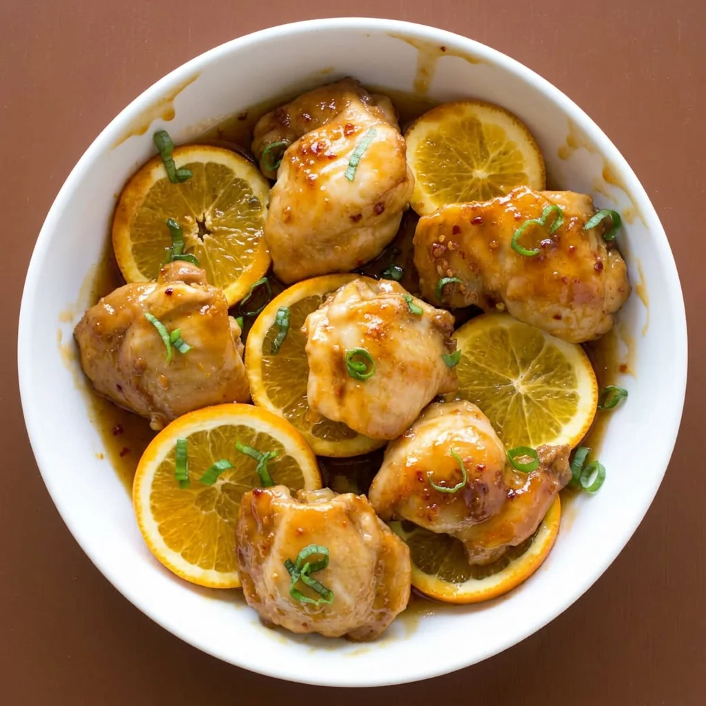 Overhead view of orange ginger glazed chicken thighs with orange slices in a white bowl